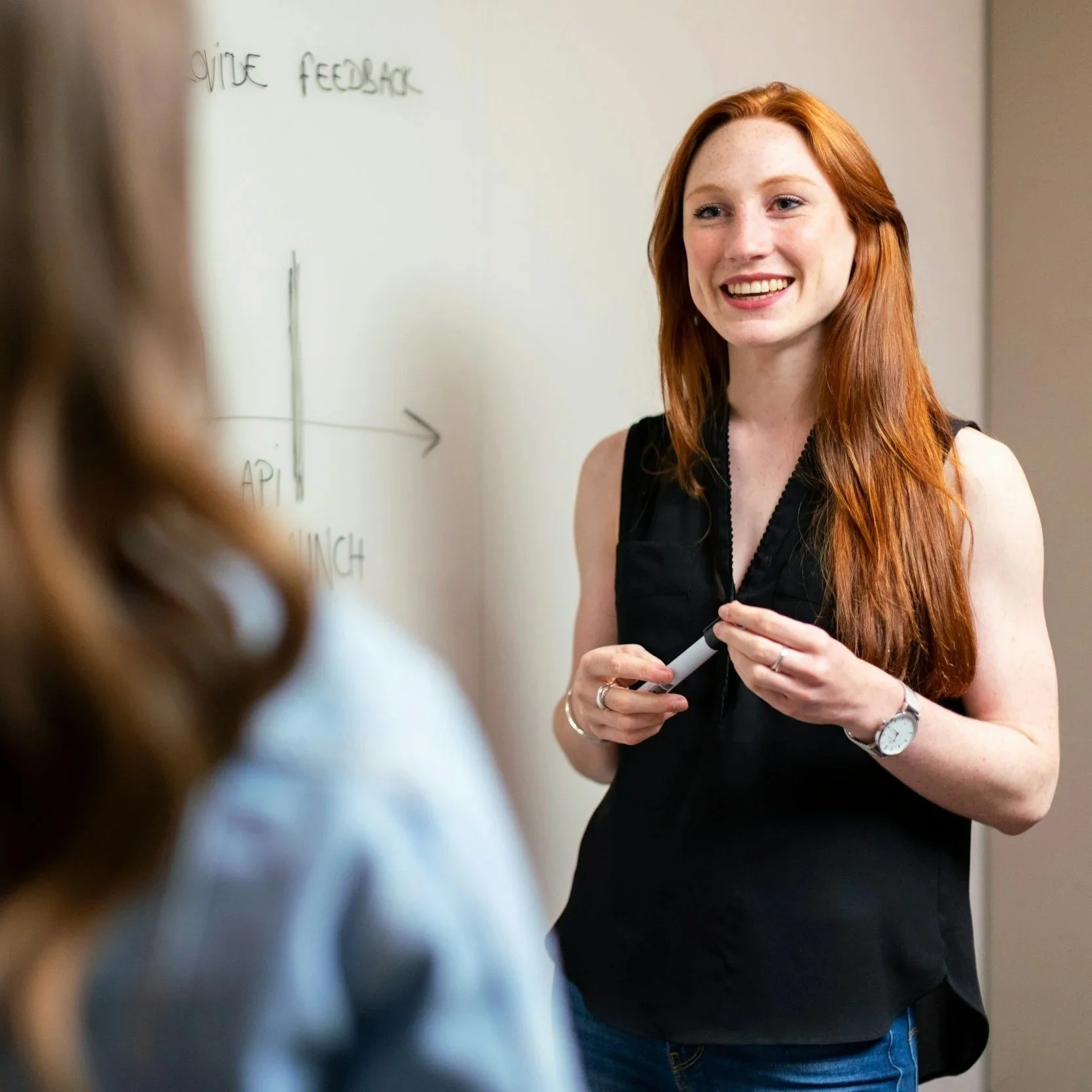 Une femme avec des cheveux roux souriante, tenant un marqueur, donnant une présentation ou un cours devant un tableau blanc.