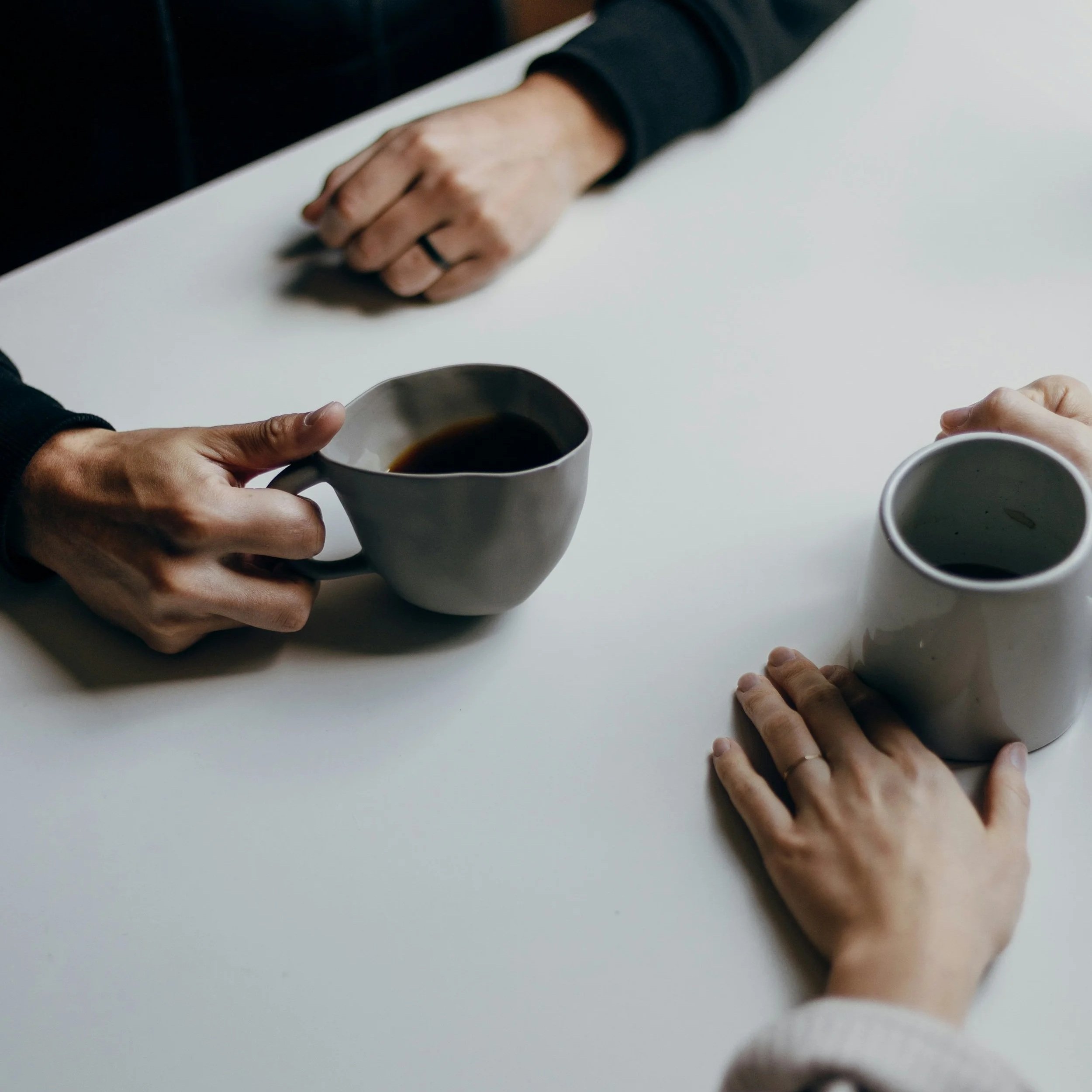 Deux mains tenant des tasses de café sur une table blanche, une main porte une tasse de café noir, l'autre repose une main avec une bague.