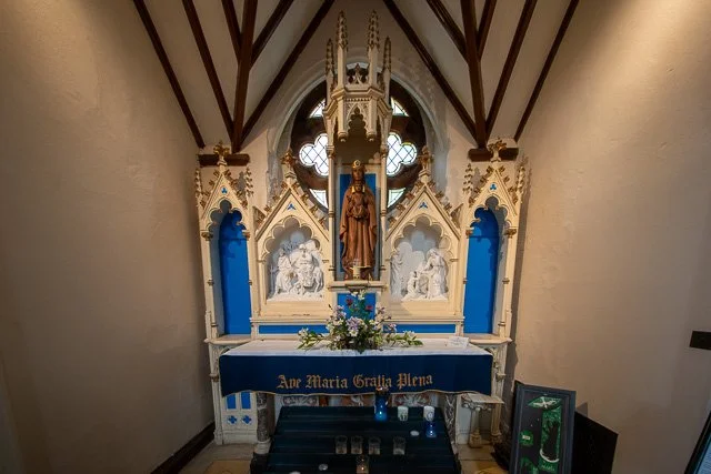 A Gothic-style altar inside a small chapel with a statue of a saint, decorative archways, and stained glass windows, adorned with flowers and religious items.