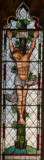 Stained glass depiction of Jesus Christ on the cross with a crown of thorns, surrounded by decorative patterns and greenery.