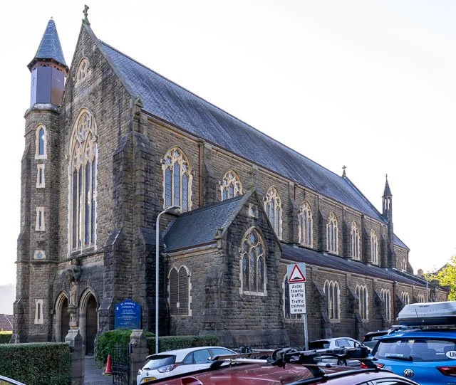 A large historic stone church with stained glass windows, a steeply pitched roof, and small towers at the corners, situated on a street with parked cars.