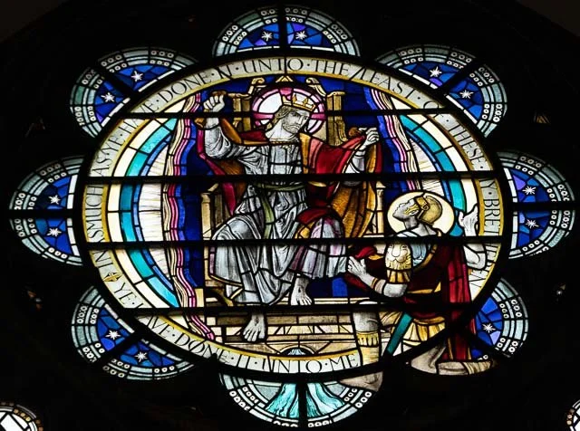Stained glass window depicting Jesus Christ on the cross with a person kneeling before him, surrounded by decorative blue and white floral patterns.