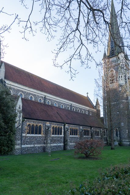 A historic stone church with a tall steeple and arched windows, surrounded by grass, trees, and shrubs.