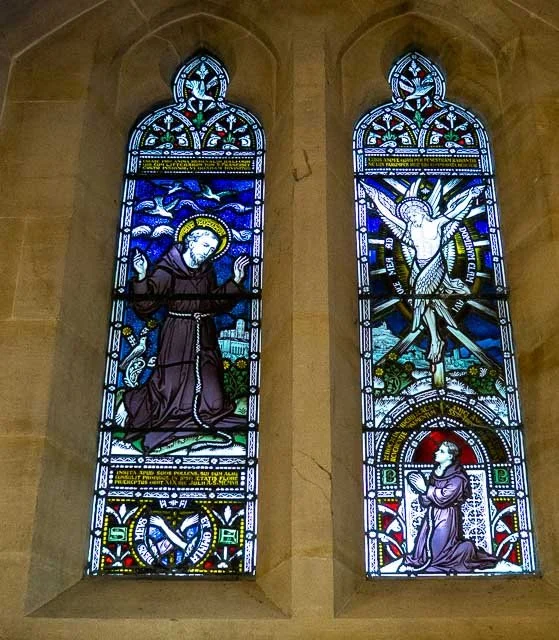 Two stained glass windows depicting religious scenes: the left shows a kneeling figure with a halo, possibly a saint, praying or in worship; the right shows a person kneeling in prayer with a depiction of the crucifixion above.