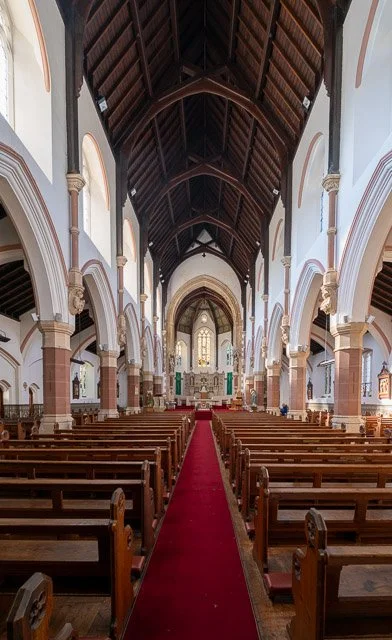 Interior of a church with wooden pews, a red carpet aisle, white walls, arched windows, and a high wooden ceiling.