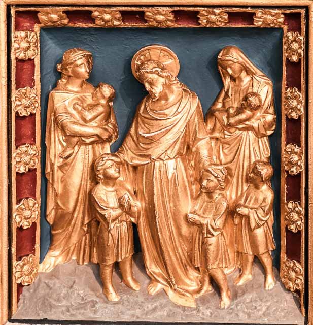 Metallic religious relief sculpture depicting Jesus Christ with children and women, surrounded by a decorated frame with floral motifs.