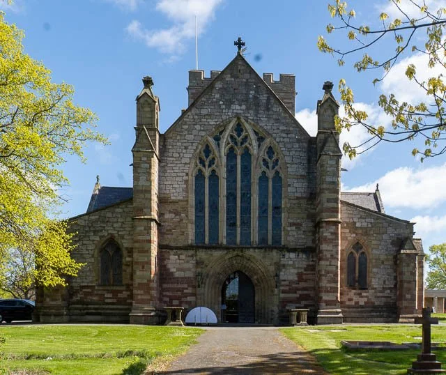Front view of a historic stone church with pointed arched windows and Gothic architectural details, surrounded by trees and grass.