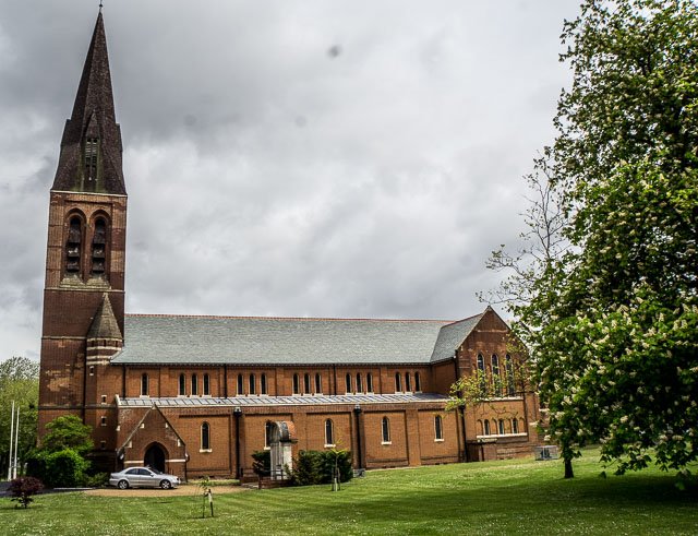 A brick church with a tall steeple, located in a grassy area with trees and parked cars nearby, under a cloudy sky.