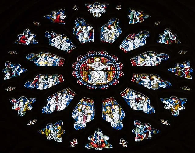 Circular stained glass window depicting religious scenes with a central figure on a throne surrounded by angels and saints.