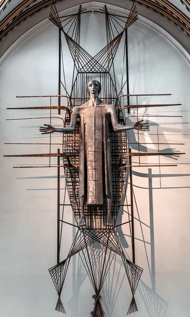 Sculpture of a human figure with outstretched arms, integrated with geometric metal structures and wires, mounted on a wall in an arched alcove.