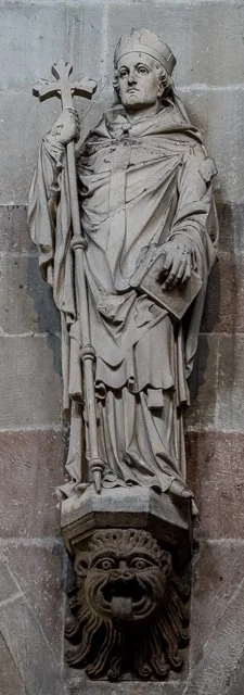 Stone statue of a crowned figure holding a staff and book, standing on a detailed lion's head base, on a wall.