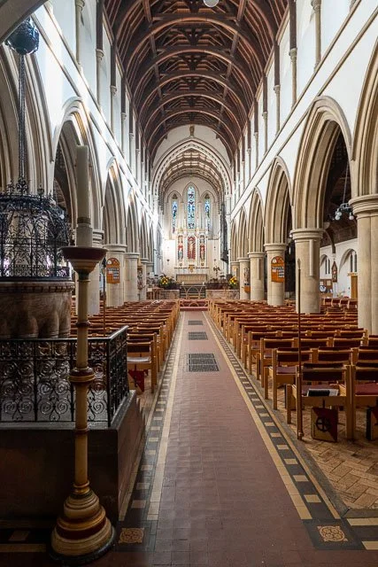 Interior view of a church with high arched wooden ceiling, columns, wooden pews, and stained glass windows at the front.
