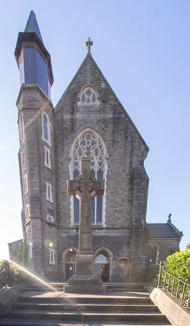 Stone church with a steeple and arched windows, situated on a hilltop with a cross on top, and green bushes nearby.
