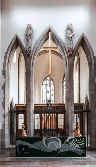 Inside a church with tall stained glass windows, stone arches, and a wooden cross above the altar, which has a decorative panel with abstract art.
