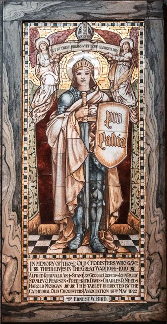 Stained glass window of a warrior saint holding a shield with the words 'Pax Patria.' Two angels hold a banner above, inscribed with the phrase 'To a true peace savior of the glorious day.' The window includes a memorial inscription for those who gav