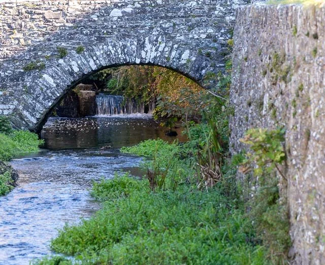 Stone bridge arch over a small creek with lush greenery and a waterfall in the background.