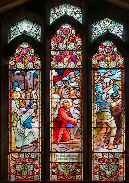 Three stained glass windows depicting a religious scene with Jesus kneeling with a crown of thorns and soldiers surrounding him, with floral and geometric patterns.
