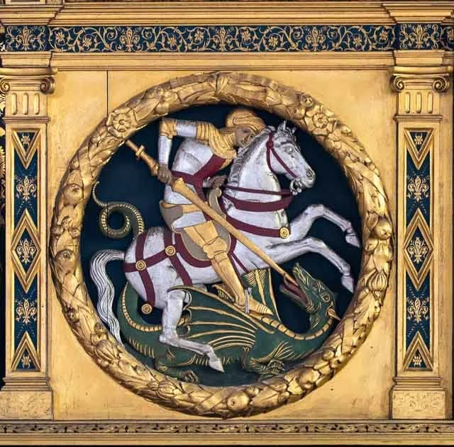 A detailed, gold-colored circular sculpture depicting a knight in armor riding a horse, holding a spear, surrounded by ornate decorative borders.