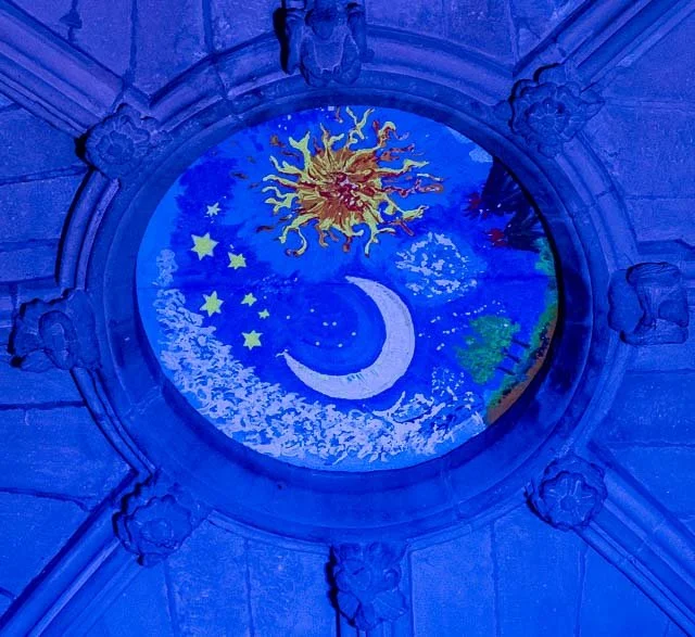 Colorful painting of a sun, moon, and stars on a circular stained glass window with a stone frame.