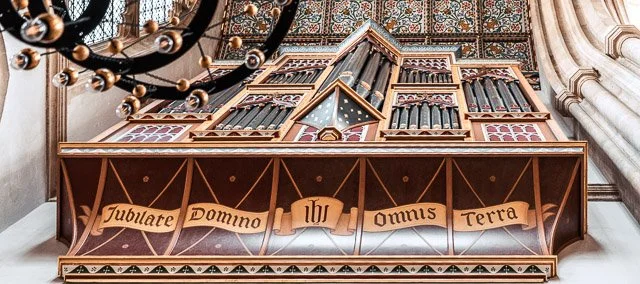 An ornate wooden pipe organ in a church or cathedral, with decorative carvings and a banner that reads 'Jubilate Domino IHU OMNIS TERRA'.