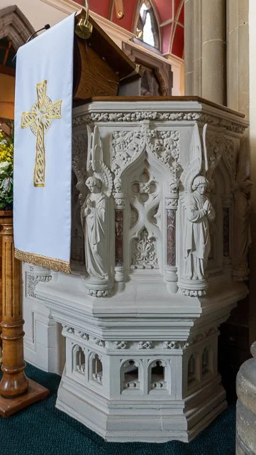 A white ornate altar with carved figures and decorative elements, located inside a church, with a wooden pulpit draped with a white cloth embroidered with a gold cross.