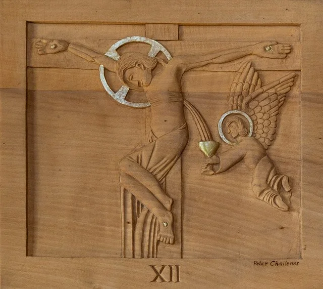 Wooden relief depicting Jesus Christ on the cross with a halo, an angel offering a chalice, and a Roman numeral XII at the bottom.