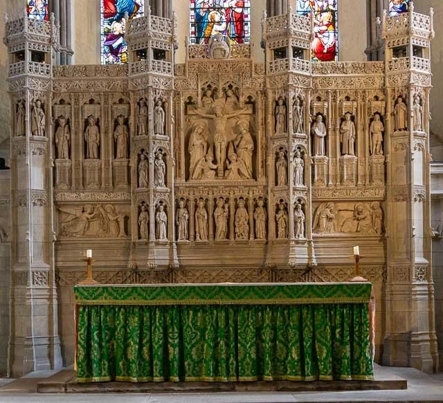 Stone altar with green embroidered cloth in front of an intricate stone religious relief sculpture, with stained glass windows above.