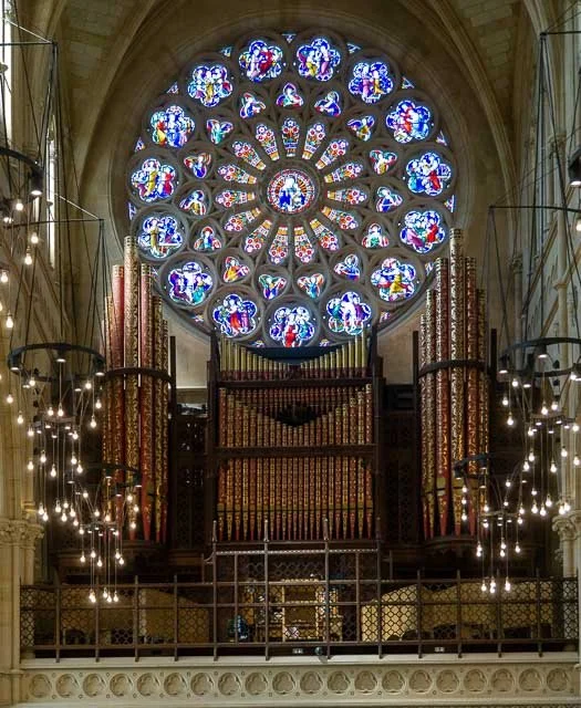 Interior of a church with a large stained glass rose window, ornate pipe organ, and decorative lighting.
