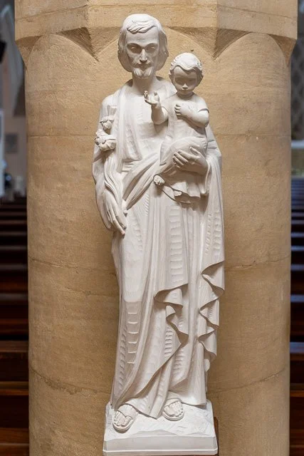 White religious statue of a man holding a child, with both figures holding what appears to be a flower, standing on a pedestal against a beige wall.