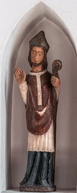 Wooden statue of a man in religious attire standing in a recessed niche, holding a staff, wearing a hat, with a hand raised in blessing.