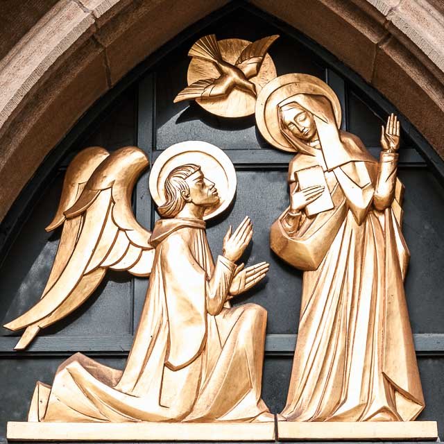 Wooden religious sculpture depicting the Annunciation, with the angel kneeling and speaking to Mary, who is holding a book and listening attentively, both with halos, and a dove above them representing the Holy Spirit.
