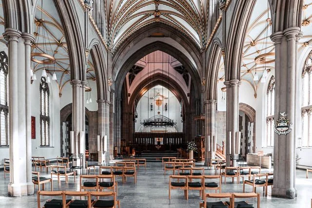 The interior of a church with high vaulted ceilings, stone columns, arched windows, and rows of wooden chairs arranged facing an altar.