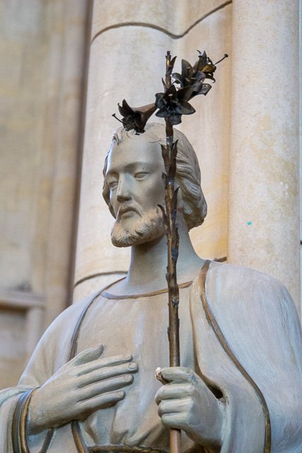 Statue of Jesus holding a staff with blackened leaves or flowers, looking contemplative, set against stone columns.