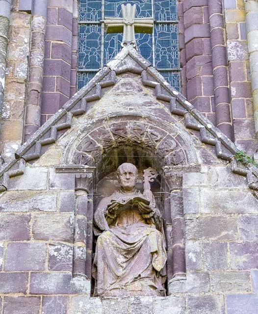 Stone sculpture of a seated religious figure, possibly a saint, inside a niche on the exterior of a church with a Gothic architectural style.