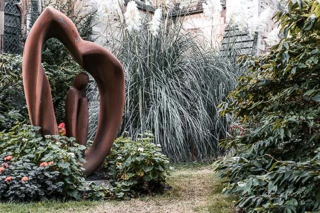 Sculpture of two abstract human figures in a garden with tall grasses and bushes.