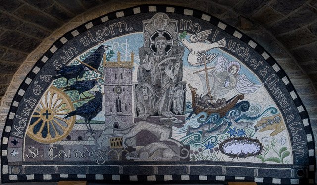 Colorful church mosaic depicting a bishop or saint seated on a throne, surrounded by symbols of Christianity, a castle, birds, and a ship on the sea, with a circular inscription around the edge.