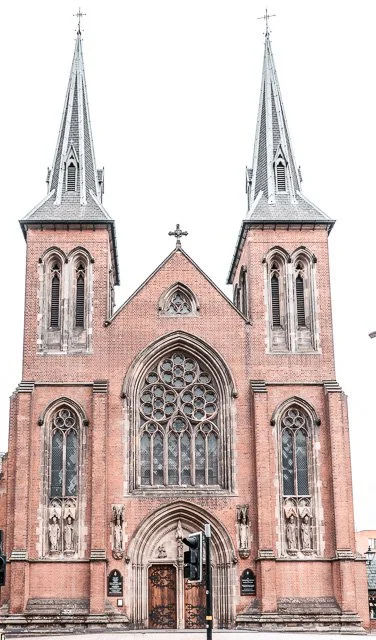 A large Gothic-style church with two tall steeples, stained glass windows, and a cross at the top, made of red brick and gray stone accents.