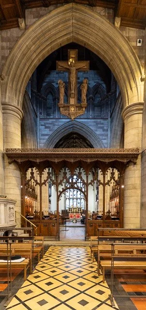 Interior view of a church with wooden pews, a patterned tile floor, and an ornate wooden altar. The background features stained glass windows, stone arches, and a large crucifix hanging from the ceiling.