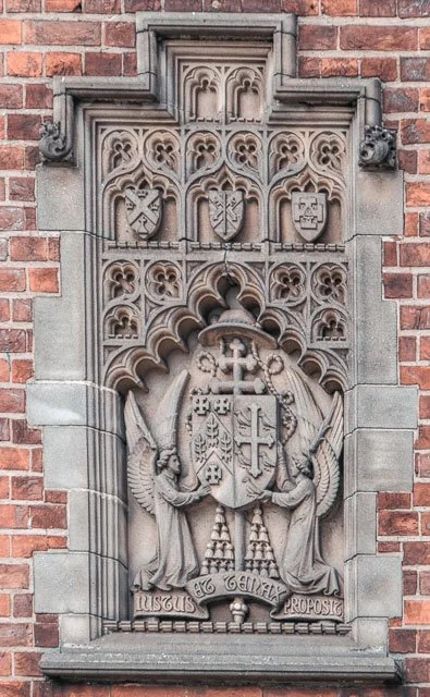 Stone carving featuring two angels holding a shield with a cross and smaller shields, floral and gothic patterns, and Latin inscriptions, mounted on a brick wall.
