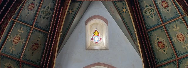 Interior of a church ceiling with ornate, patterned panels and a small stained glass window