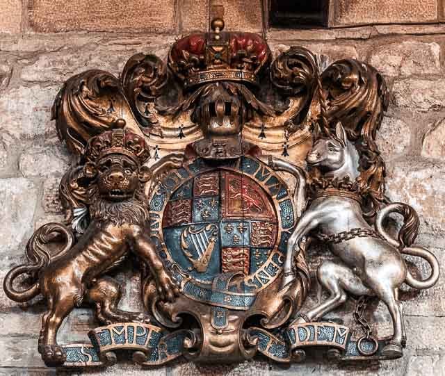 A decorative coat of arms with a lion on the left, a unicorn on the right, a crown at the top, and a shield in the center featuring the Scottish flag and symbols, mounted on a brick wall.