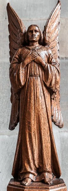 Wooden statue of an angel with large wings, standing upright and holding its hands over its chest.