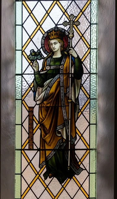 Stained glass window depicting a crowned female saint holding a bird in one hand and a staff with a cross in the other, with decorative geometric patterns in the background.