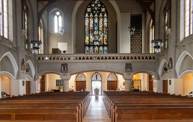 Interior of a church with wooden pews, stained glass windows, and an altar with a baptismal font at the front.