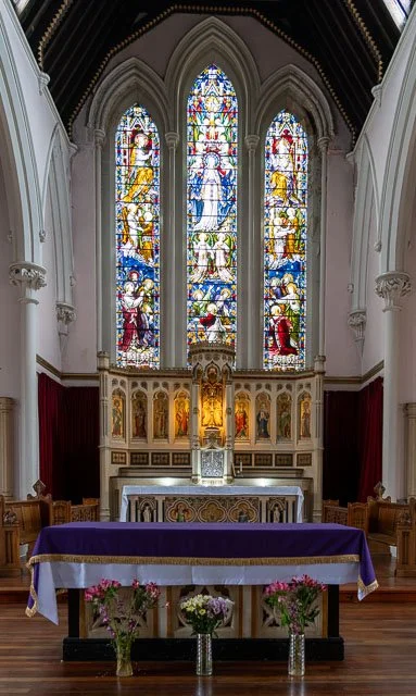 Inside a church, a stained glass window behind the altar depicting religious figures, with an ornate altar table covered with a purple cloth and flowers arranged in vases in front.