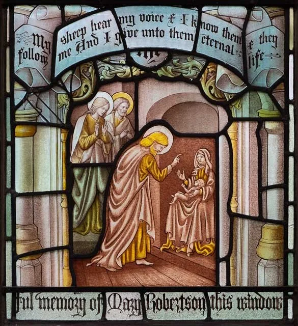 Stained glass window depicting a biblical scene with Jesus speaking to a seated woman, with three figures observing from behind. The window has decorative arches and a Latin inscription at the top.