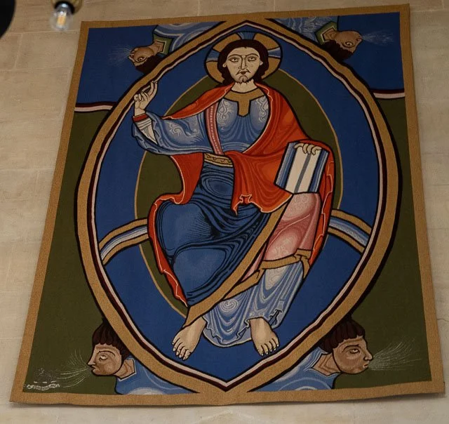 Artwork of Jesus Christ seated with a book in his left hand, blessing with his right hand, surrounded by a circular frame with two faces at the bottom, set against a green and blue background.