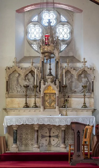 Ornate church altar with religious statues, candlesticks, and a central icon, topped by stained glass windows in a historic chapel.