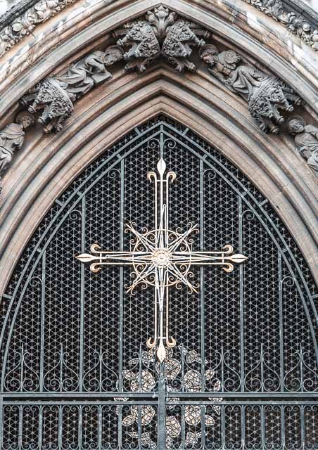 Close-up of a large, ornate clock with intricate metalwork, part of a historic building's facade with detailed stone carvings and gothic architectural elements.