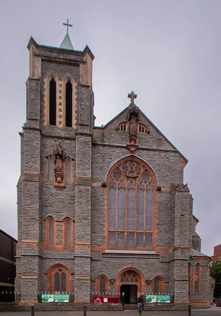 Front view of a historic stone church with a tall bell tower, arched stained glass windows, and a cross on top, under an overcast sky.